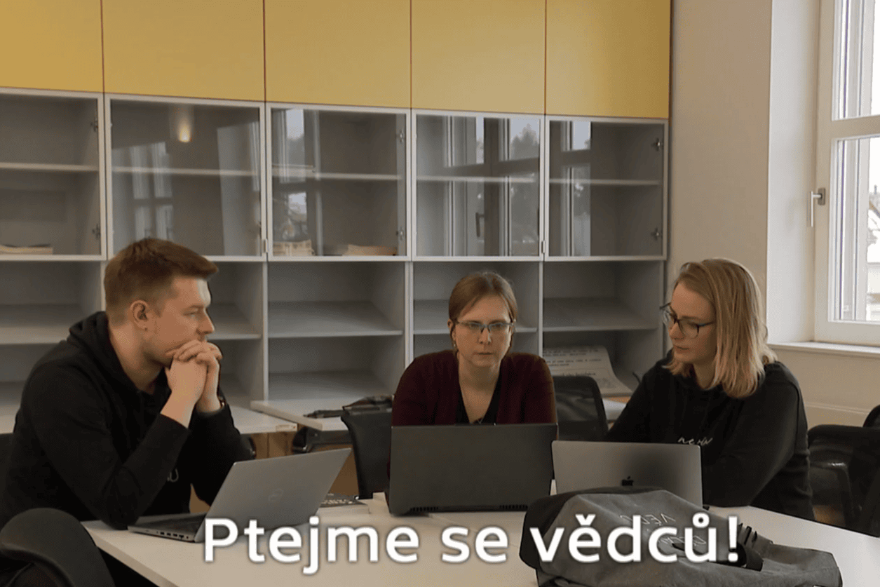 tři vědci sedí u stolu s notebooky, nápis: Ptejme se vědců